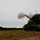 A self-propelled howitzer, consistent with an M109 Paladin, is captured mid-fire on an open training field. A projectile is visibly airborne above the muzzle, accompanied by a large smoke discharge from the barrel. The setting appears to be a military training range surrounded by a treeline under overcast skies.