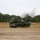 A self-propelled howitzer, consistent with the M109 Paladin series, is captured mid-fire on an open sandy training range. A projectile is visibly airborne above the muzzle, accompanied by a large white smoke plume from the discharge. The setting appears to be a military training facility surrounded by pine trees.