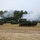 Three self-propelled howitzers, identifiable as M109 Paladin variants, are firing simultaneously in a field exercise. Significant white smoke is visible emanating from the muzzles and surrounding the vehicles, indicating active discharge. The vehicles are positioned in a line formation on an open, dry grass field with a tree line in the background.