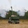 A self-propelled howitzer, consistent with the M109 Paladin series, is photographed from the rear in a field environment immediately after firing. A large plume of white and gray smoke billows from the muzzle brake and surrounds the vehicle, indicating a recent or ongoing discharge. The setting appears to be a military training range with open grassland and a treeline in the background.