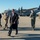 C-130J Super Hercules aircraft debuts at Kentucky Air Guard A group of civilians and military personnel are gathered on an airfield tarmac in front of a U.S. Air Force C-130 Hercules transport aircraft. A man in a long black coat and sunglasses walks prominently in the foreground, accompanied by other civilians and uniformed service members. The setting appears to be an official visit or military event at a National Guard or Air Force base.