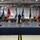 Kentucky Air Guard enters new era with arrival of C-130J Super Hercules aircraft A formal military ceremony is taking place inside a large hangar or drill hall, with a speaker standing at a podium on a raised stage draped in navy blue. Multiple flags are displayed on the stage, including the United States flag, a state flag, and several military branch/unit flags. An audience of uniformed military personnel and civilians, many wearing face masks, are seated in rows facing the stage.