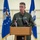 Kentucky Air Guard enters new era with arrival of C-130J Super Hercules aircraft A U.S. Air Force officer identified by his name tag as Bruce Bancroft is speaking at a wooden podium equipped with multiple microphones. He is wearing an olive green flight suit with unit patches and a name tag, standing before two blue U.S. Air Force flags in what appears to be a hangar or official military facility. The setting suggests a formal press briefing or official military ceremony.