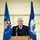 Kentucky Air Guard enters new era with arrival of C-130J Super Hercules aircraft An elderly man with white hair and glasses stands at a wooden podium with two microphones, speaking at a formal event. He is dressed in a dark blazer over a burgundy argyle sweater vest. Two blue ceremonial flags flank him on either side, one of which is partially readable as an 'Air National Guard' flag.
