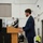 Kentucky Air Guard enters new era with arrival of C-130J Super Hercules aircraft A young man in a navy blue suit is delivering a speech at a wooden podium equipped with two microphones. The setting appears to be a large industrial or institutional facility, such as a hangar or warehouse, with white walls and high ceilings. At least one other person is visible in the background wearing a face mask.