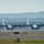Kentucky Air Guard enters new era with arrival of C-130J Super Hercules aircraft Two military turboprop transport aircraft, consistent with C-130 Hercules variants, are taxiing or positioned on an airfield tarmac. The aircraft appear in gray military livery with all four propeller engines running, visible from heat distortion effects. A large industrial or hangar building is visible in the background, with tree-covered hills beyond.