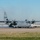 Kentucky Air Guard enters new era with arrival of C-130J Super Hercules aircraft A U.S. Air Force C-130 Hercules military transport aircraft is taxiing on an airfield tarmac with all four turboprop engines running, producing visible exhaust haze. A ground crew member in military uniform is marshaling the aircraft using hand signals. An airport fire/rescue truck labeled 'R-64' is positioned nearby in a standby capacity.