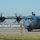 Kentucky Air Guard enters new era with arrival of C-130J Super Hercules aircraft A military C-130 Hercules turboprop transport aircraft is taxiing on a runway while a ground crew member in camouflage uniform directs it using hand signals. All four propellers are spinning, visible through motion blur. A flag is mounted on top of the fuselage near the cockpit, and the aircraft bears military gray livery.