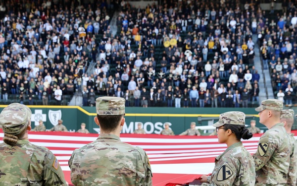 U.S. Army soldiers in OCP (Operational Camouflage Pattern) uniforms are unfolding or displaying a large American flag on a baseball field during a pre-game or ceremonial event. The stadium is filled with a large crowd of spectators standing in the background. A military vehicle is visible in the right background, suggesting a military appreciation or patriotic ceremony.