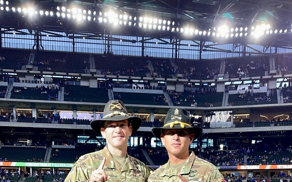 Two U.S. Army soldiers in Operational Camouflage Pattern (OCP) uniforms and cavalry Stetson hats pose on the field of an indoor NFL stadium, both making a 'number one' finger gesture. The setting appears to be a military appreciation event at what looks like AT&amp;T Stadium or a similar modern domed football venue. Players can be seen warming up in the background on the well-lit artificial turf field.