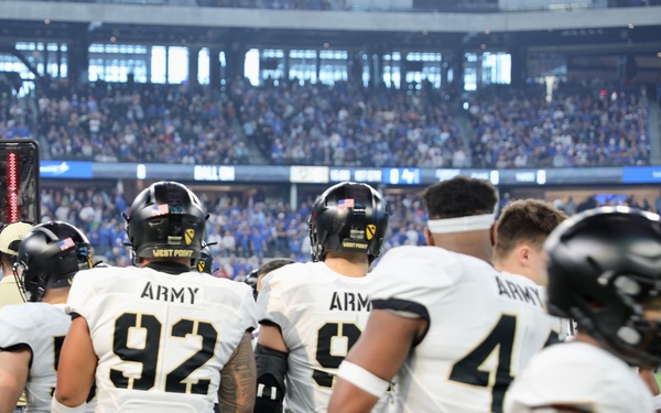 The image shows Army West Point Black Knights football players standing on the sideline during a game played in a large indoor stadium. Players are wearing white jerseys with black 'ARMY' lettering and black helmets bearing the '1st Cavalry Division' patch and 'WEST POINT' text. The stadium appears to be filled with spectators, suggesting a significant game event.
