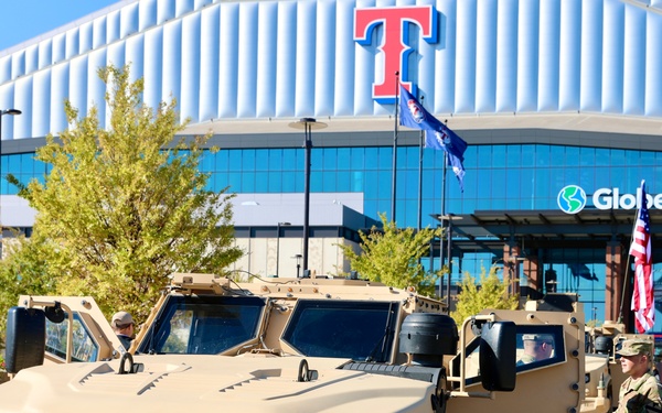 Military vehicles, identified as Joint Light Tactical Vehicles (JLTVs), are parked in front of Globe Life Field, home of the Texas Rangers MLB team in Arlington, Texas. Soldiers in camouflage uniforms are present alongside the tan-colored armored vehicles. This appears to be a military display or ceremonial event at the stadium.