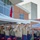 A formal United States Marine Corps ceremony is taking place outdoors, likely a change of command or official military event. Multiple Marines in dress uniform are rendering salutes simultaneously, with senior officers prominently positioned in the foreground. The setting includes a tent canopy, red ceremonial chairs bearing the USMC eagle globe and anchor emblem, and a red flag consistent with a general officer's flag.