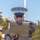 A United States Marine in dress uniform (Charlie uniform) holds an ornamental scroll and a ceremonial Mameluke sword during what appears to be a formal military ceremony. The Marine is wearing the distinctive white cover (cap) with the Eagle, Globe, and Anchor emblem, along with a khaki service shirt and blue trousers. Additional Marines in similar uniforms are visible in the background.
