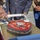 A military cake-cutting ceremony is taking place outdoors, featuring a shield-shaped cake decorated in red and gray with a military unit insignia and the partial text 'OVE YOU NEVER BELOW YOU' visible around the base. Personnel in U.S. Marine Corps uniforms (tan service uniforms with red-striped blue trousers) and a civilian in a blue suit are gathered around the cake. A sword or ceremonial knife is being used to cut the cake, consistent with traditional Marine Corps Birthday Ball or unit celebration customs.