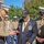 A military ceremony is taking place outdoors, likely at a U.S. Marine Corps installation. A senior Marine officer in dress uniform is presenting something on a red plate to an elderly veteran wearing a Purple Heart cap and a navy blazer adorned with multiple medals and a medallion. Additional Marines in service uniforms stand in formation in the background.