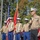 A senior U.S. Marine Corps officer, identifiable by his white cover, multiple service ribbons, and general officer rank insignia, addresses an audience via microphone at an outdoor military ceremony. Behind him stands a color guard of four Marines in dress uniform holding the U.S. flag, Marine Corps colors, and additional unit guidons. The setting appears to be a well-lit outdoor venue with trees in the background, suggesting a formal military event such as a change of command or commemoration ceremony.