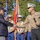 A formal military ceremony is taking place outdoors, where a senior U.S. Marine Corps officer in service uniform is shaking hands with an elderly civilian veteran wearing a Purple Heart cap. Color guard Marines in dress blues stand at attention in the background holding flags including the U.S. flag. The event appears to be a recognition or commemoration ceremony.