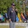 An elderly veteran is speaking into a microphone at an outdoor ceremony, holding a cane in his left hand. He is wearing a navy blazer with a visible organizational patch, a medal/lanyard, and a black cap with purple lettering that reads 'Purple Heart' and 'Combat Wounded'. Multiple U.S. state flags are displayed on poles in the background.