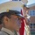 A close-up profile shot of a young female Marine or ROTC cadet in dress uniform, wearing a white cover (cap) with gold eagle globe and anchor insignia and a black patent leather visor. She stands at attention alongside at least one other uniformed individual in the background, with an American flag visible and a red brick building behind them. The image appears to capture a formal military ceremony or parade formation.