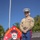 A U.S. Marine in Service 'Charlie' uniform stands at parade rest beside a bulldog on a leash. Behind them is a unit insignia sign reading 'Swift Silent Deadly – 2D Recon Battalion' featuring a skull-and-crossbones design on a red background. The setting appears to be an outdoor military installation on a clear, sunny day.