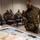 Concept to capability: Naval Warfighting Symposium A military briefing or planning session is underway in an indoor facility, with personnel in camouflage uniforms gathered around a large map spread on a table. A senior officer, identifiable by a star insignia on his uniform, is examining the map closely while others observe from seated positions. All personnel are wearing black face masks, suggesting this was taken during the COVID-19 pandemic period.