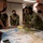 Concept to capability: Naval Warfighting Symposium A group of military personnel wearing camouflage uniforms and black face masks are gathered around a table reviewing large maps and documents. One individual appears to be briefing or pointing out details on the maps to the group. The setting appears to be an indoor military planning or operations room.
