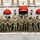 Concept to capability: Naval Warfighting Symposium A large group of military personnel, consisting of both U.S. Marines and U.S. Navy sailors, pose for a formal group photograph in front of Julian C. Smith Hall, headquarters of II Marine Expeditionary Force. The personnel are dressed in camouflage utility uniforms and are arranged in a single row spanning the width of the frame. The building features distinctive red awnings with 'II Marine Expeditionary Force' lettering over the main entrance.