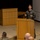 Concept to capability: Naval Warfighting Symposium A military officer in olive green flight suit is delivering a presentation or briefing at a wooden podium to an audience of seated military personnel. The speaker appears to be gesturing with both hands while addressing the group in what appears to be a formal briefing room or auditorium. Several audience members in military uniforms are visible from behind in the foreground.