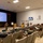 Concept to capability: Naval Warfighting Symposium A military briefing is taking place in an auditorium-style conference room where a presenter in camouflage uniform is delivering a 'Naval Logistics Primer' presentation to an audience of military personnel. The attendees, predominantly in USMC digital camouflage uniforms, are seated in rows of theater-style chairs. A secondary monitor on the side wall suggests a hybrid in-person and remote attendance format.
