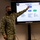 Concept to capability: Naval Warfighting Symposium A U.S. Navy service member wearing a black face mask and NWU Type III camouflage uniform is pointing at a Sony display screen showing a 'Friendly Force (Traditional)' organizational chart. The briefing appears to take place in a military simulation or training center, as indicated by text visible on the monitor stand. The chart displays a command hierarchy including CTF 950, CTG 950.2, and CTG 950.3 with various naval vessels and units listed beneath.