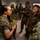 Concept to capability: Naval Warfighting Symposium A group of military personnel in camouflage uniforms are gathered in what appears to be an indoor briefing or meeting area. A female service member in U.S. Marine Corps MARPAT camouflage is speaking and holding a green notebook, appearing to brief or converse with colleagues. The group includes personnel wearing different camouflage patterns, suggesting a multinational or multi-branch gathering.
