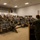 Concept to capability: Naval Warfighting Symposium A military briefing or training session is taking place in an auditorium-style room, with a uniformed speaker addressing an audience of approximately 30-40 military personnel seated in tiered rows. All individuals are wearing face masks and camouflage uniforms consistent with U.S. Navy NWU Type III pattern. Seats marked with red X markers indicate social distancing protocols are in effect.