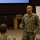 Concept to capability: Naval Warfighting Symposium A U.S. Navy officer in NWU Type III camouflage uniform is addressing a group of military personnel in what appears to be a briefing or training session. The speaker's name tape reads 'MENONI' and he wears a U.S. Navy breast insignia. A whiteboard with handwritten notes and a projection screen are visible in the background.