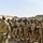 A military training or briefing session is taking place in an outdoor area surrounded by damaged or deteriorating buildings. One instructor in MultiCam/OCP pattern camouflage is addressing a group of soldiers wearing desert camouflage uniforms and carrying rifles. A civilian interpreter in a green shirt appears to be facilitating communication between the instructor and the group.