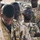The image shows a group of military personnel wearing desert camouflage uniforms gathered together, likely during a briefing, formation, or operational pause. At least one soldier wears reflective sunglasses and has crossed arms, while others appear attentive or looking downward. A military vehicle is partially visible in the background.