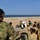 A military training or briefing session is taking place on a sandy coastal beach environment. A person in multicam camouflage uniform and black cap observes from an elevated position near a vehicle, while a group of soldiers in desert tan uniforms and helmets attend a presentation at a whiteboard. The setting appears to be an outdoor field instruction scenario near an ocean shoreline.