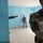 The image shows multiple individuals in military camouflage uniforms inside a building with blue-painted walls. The foreground figure wears a US flag patch on the right shoulder and has visible tattoos on the forearm. Personnel appear to be conducting an indoor clearing or assessment exercise.
