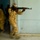 A soldier in desert camouflage uniform is photographed mid-movement aiming an AK-pattern rifle in what appears to be an indoor training or clearing exercise. Additional personnel in similar uniforms are visible in the blurred background moving through a doorway. The motion blur suggests dynamic movement, consistent with a room-clearing or tactical training drill.