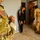 A U.S. Army soldier in MultiCam/OCP camouflage uniform is visible in the foreground, identifiable by the American flag patch on the left shoulder. In the background, what appears to be a joint operation is underway inside a building, with at least one civilian and another soldier in a different camouflage pattern visible. The scene suggests a military search, inspection, or advisory operation conducted indoors.