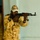 A person wearing desert camouflage uniform and a baseball cap is aiming an AK-pattern rifle directly toward the camera. Additional individuals in similar uniforms are visible in the blurred background. The setting appears to be an indoor or semi-indoor environment with a light-colored wall.