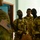 A group of soldiers wearing desert camouflage uniforms are attentively listening to a briefing or instruction from a US military personnel visible in the foreground left. The US service member is identifiable by the American flag patch on their MultiCam/OCP uniform sleeve. The setting appears to be an indoor facility, suggesting a training or coordination meeting context.