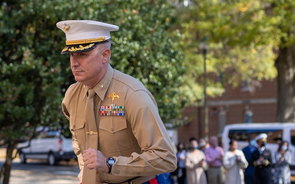 A senior U.S. Marine Corps officer in Service 'C' uniform is ascending stairs during what appears to be an official military ceremony or event. He displays multiple rows of service ribbons and general officer rank insignia, suggesting high rank. A crowd of military personnel and civilians observes in the background.
