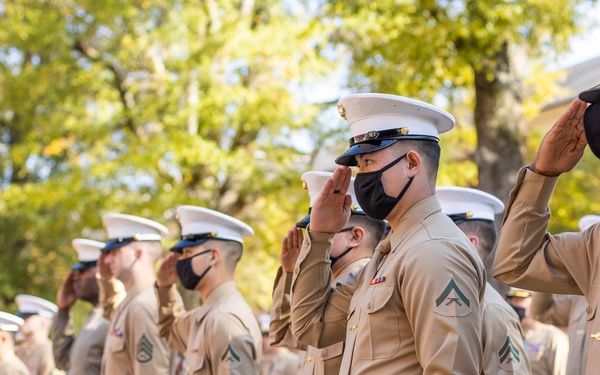 A formation of United States Marines in dress uniform are executing a salute during what appears to be a formal military ceremony or parade. The Marines are wearing the standard Service 'C' uniform consisting of khaki shirts, blue trousers with red stripe, and white covers (caps). All visible personnel are wearing black face masks, suggesting this event took place during the COVID-19 pandemic period (approximately 2020-2021).