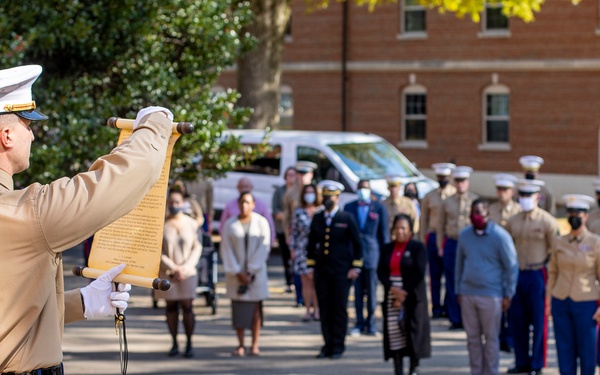 A U.S. Marine in dress uniform reads from a ceremonial scroll to a formation of military personnel and civilians gathered outdoors. The setting appears to be a formal military ceremony, likely a reading of orders, a proclamation, or a birthday message, conducted on a paved courtyard in front of a brick building. Several attendees are wearing face masks, suggesting the event took place during the COVID-19 pandemic period.