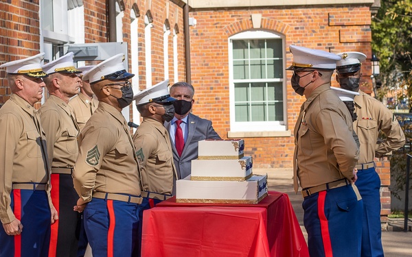 A group of U.S. Marines in dress blues and service uniforms are gathered around a tiered white and gold ceremonial cake on a red-draped table, likely for a Marine Corps Birthday celebration. A civilian in a gray suit and red tie stands behind the cake. The event appears to be an outdoor formal ceremony in front of a brick building.