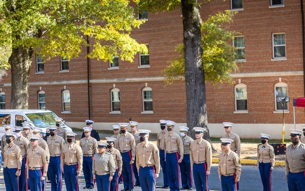 A formal U.S. Marine Corps military ceremony is taking place outdoors on a paved plaza, with a large formation of Marines in dress uniform standing at attention. Civilians and additional military personnel observe from the left side. The setting appears to be a military installation, with a red brick building and red canopy tent visible in the background.