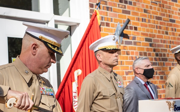 A U.S. Marine Corps officer is cutting a ceremonial cake with a military sword during what appears to be a Marine Corps Birthday celebration. Several Marines in service uniforms and a civilian in a suit stand in the background near a red Marine Corps flag. The cake is decorated with gold trim and bears the text 'Semper Fidelis' and 'Once a' (likely 'Once a Marine, Always a Marine').