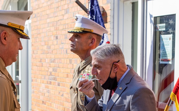 Three individuals stand outside a brick building near an American flag. Two are wearing United States Marine Corps dress uniforms, while a third man in a gray suit and lowered face mask is eating a slice of decorated cake from a white plate. The setting appears to be a formal or ceremonial occasion.