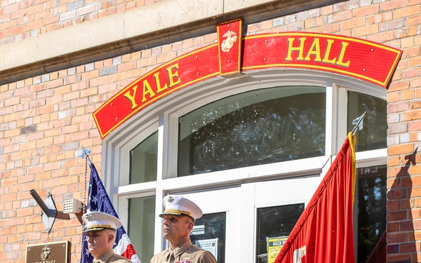 Two United States Marine Corps officers stand at attention in front of Yale Hall, a brick building marked with USMC insignia. They are in service uniform (Charlie/Delta service uniforms) flanked by the American flag and a red Marine Corps unit flag. A commemorative plaque for Yale Hall is visible on the left wall.